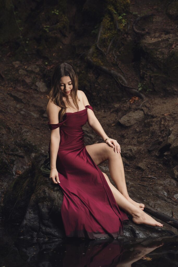 High school senior in a burgundy gown seated barefoot on the rocks beside Dickerson Creek near Bremerton