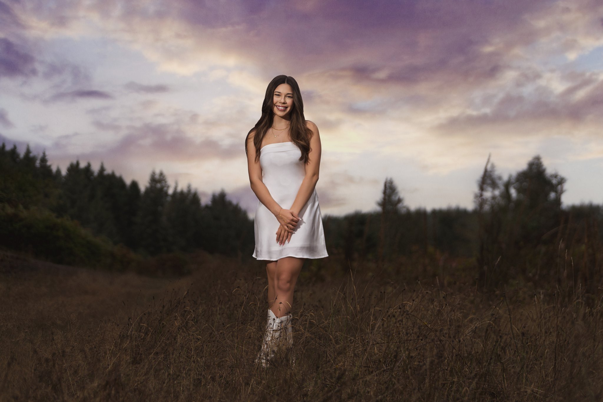 High school senior in a white dress and cowboy boots in a golden meadow under a pastel sunset sky near Bremerton. Captured by Kitsap County senior photographer Nautical Moments Photography.