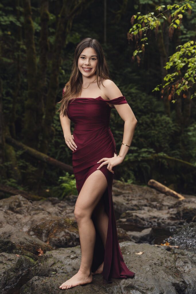 High school senior in a burgundy gown standing on creek rocks at Dickerson Falls near Bremerton