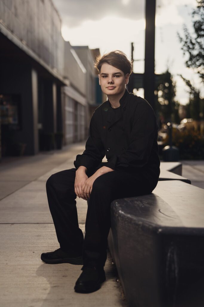 Culinary senior in a chef coat sitting on a concrete bench along 4th Street in downtown Bremerton at golden hour
