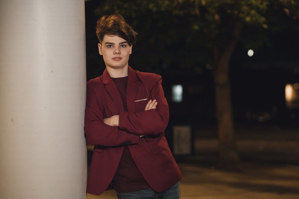 High school senior in a burgundy blazer with arms crossed leaning on a white column at night in downtown Bremerton