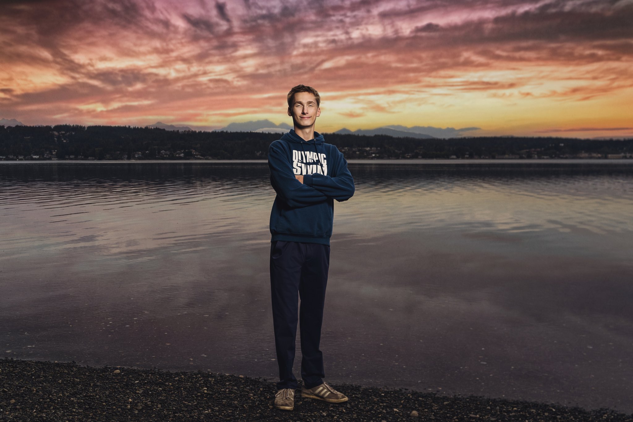 High school senior in an Olympic Swim hoodie standing on the Dyes Inlet beach at J.A. and Anna F. Smith Childrens Park in Bremerton under a blazing sunset. Photographed by Bremerton senior photographer Nautical Moments Photography.