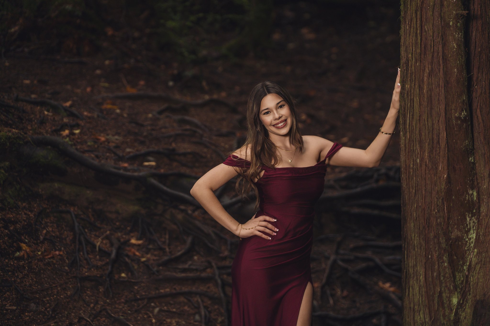 High school senior in a burgundy gown beside a mossy cedar trunk in a dark Pacific Northwest forest