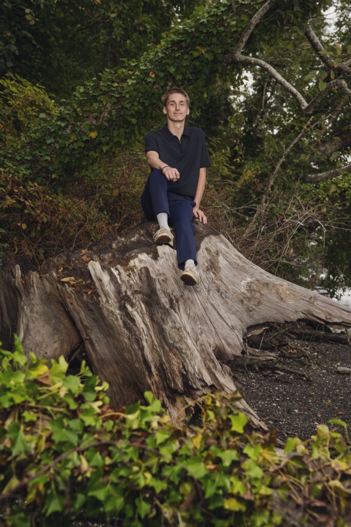 High school senior sitting on a giant driftwood stump on the Dyes Inlet beach at Smith Childrens Park