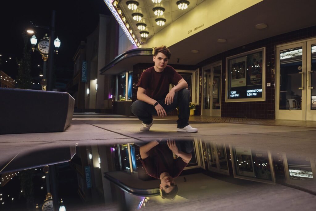 High school senior crouching beneath the historic Roxy Theatre marquee in downtown Bremerton with his reflection on the wet pavement at night