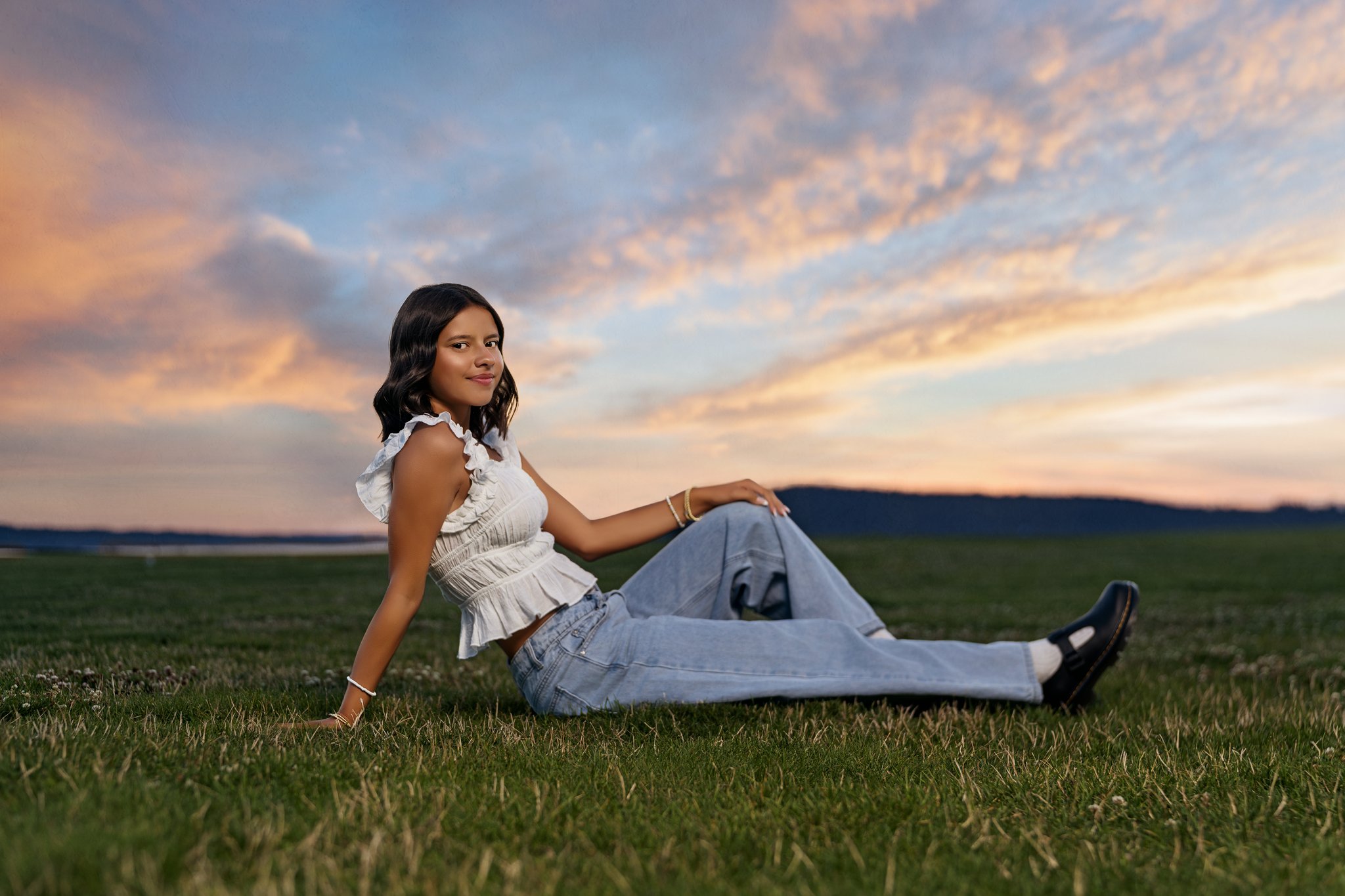 High school senior in a white ruffled top and jeans sitting on the great lawn at Chambers Bay at sunset