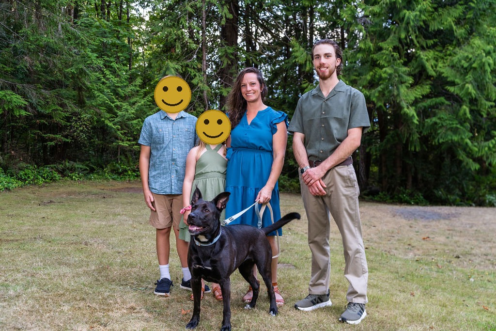 Family of four with their dog standing together in a grassy clearing, faces covered for privacy
