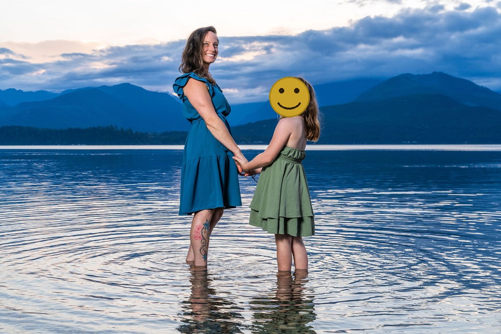 Family photo session with a mother and daughter standing in calm water at dusk in Washington, faces covered for privacy