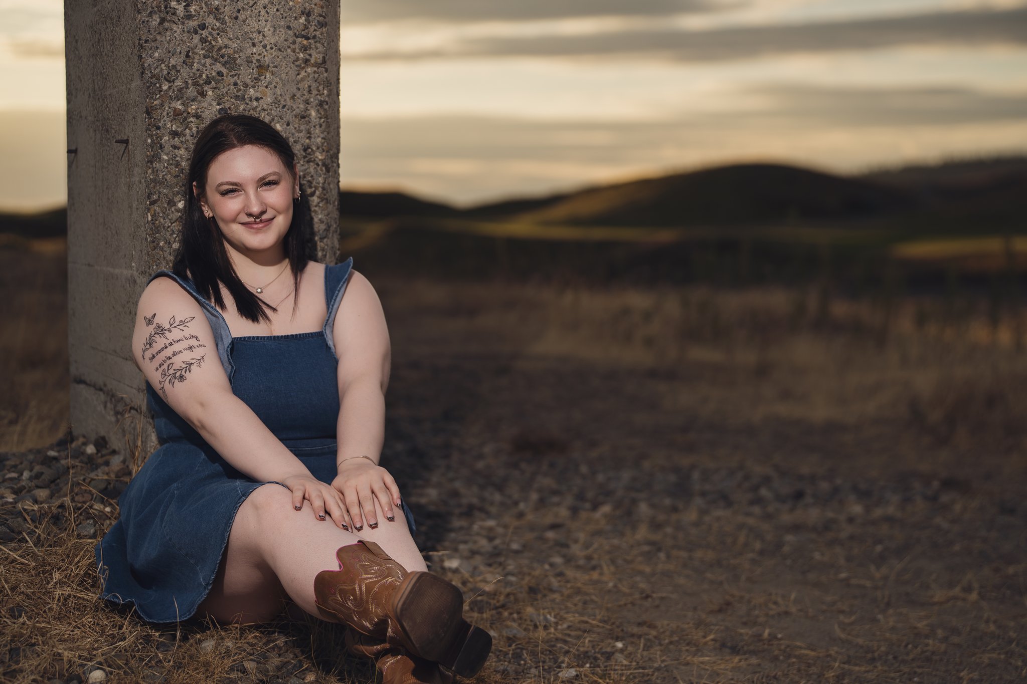 Senior in a denim dress and cowboy boots sitting at the base of a concrete column at the Chambers Creek ruins near University Place. Photographed by Nautical Moments Photography.
